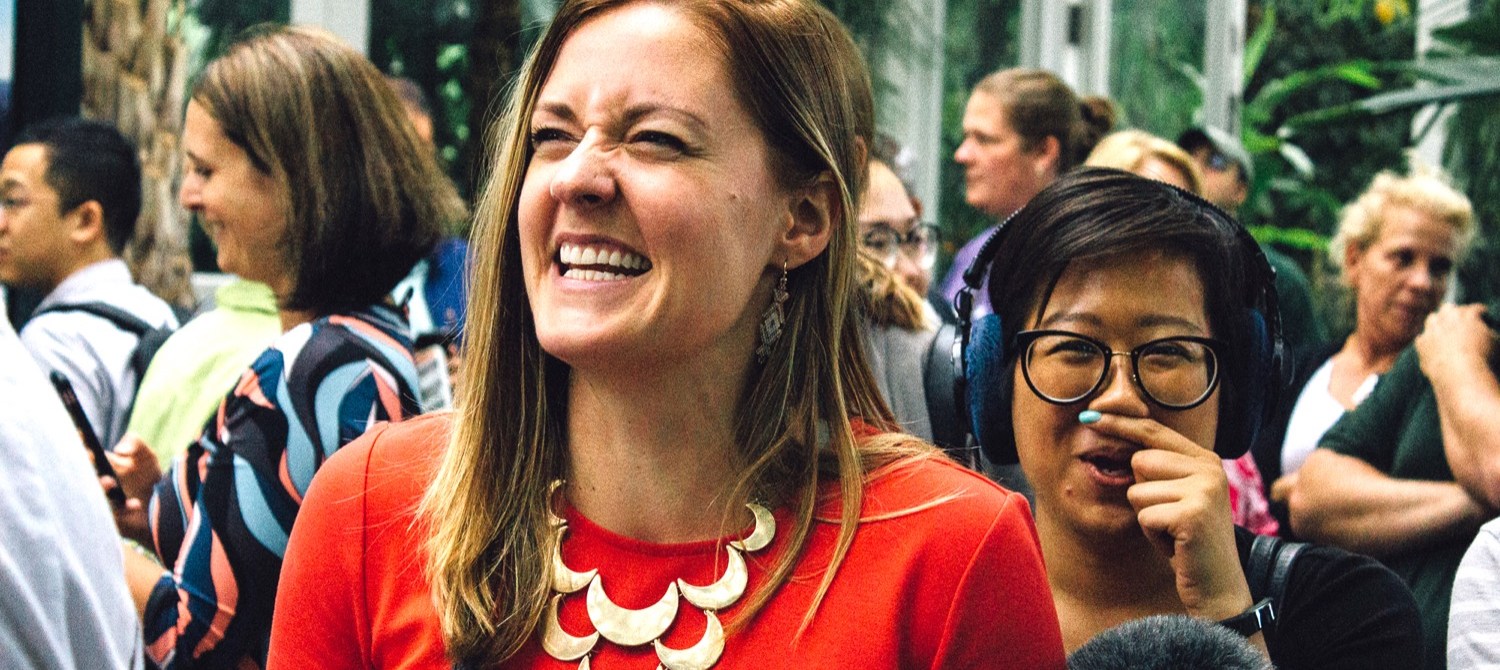 in a crowd of people, there are two women, one who's nose is wrinking and smiling, and the other, wearing headphones and carrying a microphone, is putting a finger under her nose