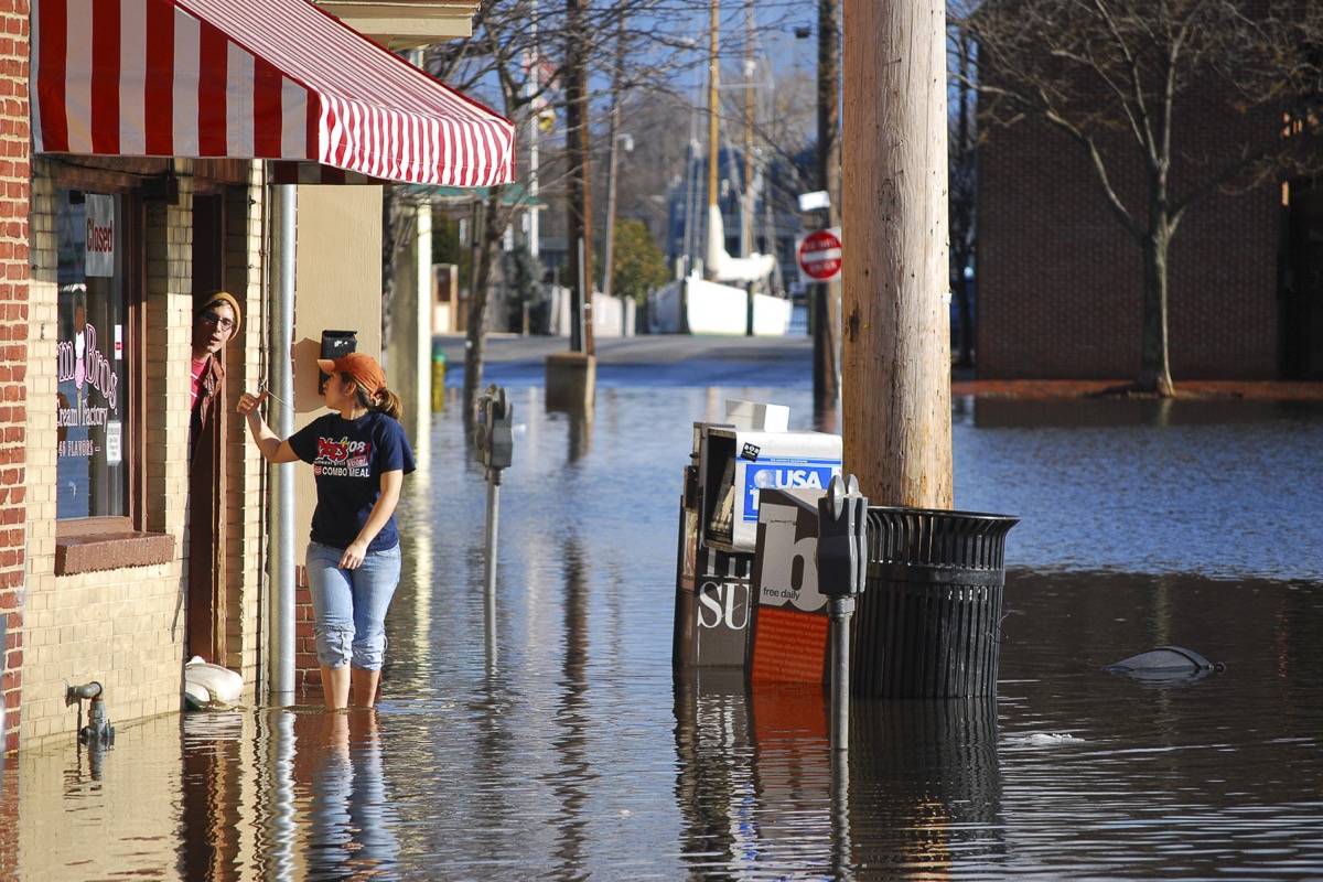 a street is flooded up to a woman's shin. she has her jeans rolled up and is waving to a man who is sticking his head out a door that is barricaded with sandbags.