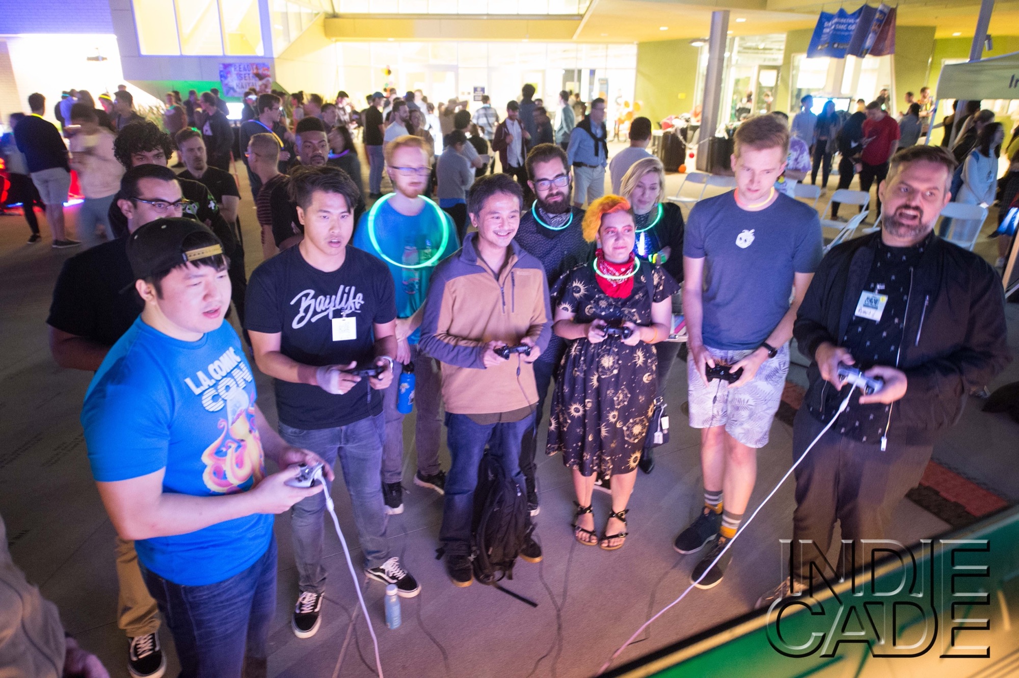 a dozen people stand in a semicircle looking at a large screen off camera. six of them hold video game controllers and a large crowd is behind them. it's at night outdoors