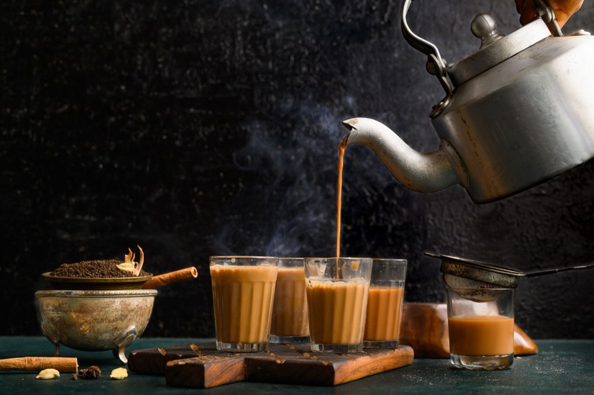 Milky brown tea is poured from a gray metal tea kettle into clear glasses surrounded by sticks of cinnamon and other spices.