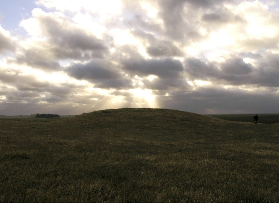 A view of a flat grassy area and a long, long hump that rises just above the horizon. Bright clouds cast rays of light on the shaded landscape.