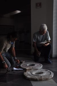 Two men couch over two large stone disks that sit on the floor, in a mostly darkened room.