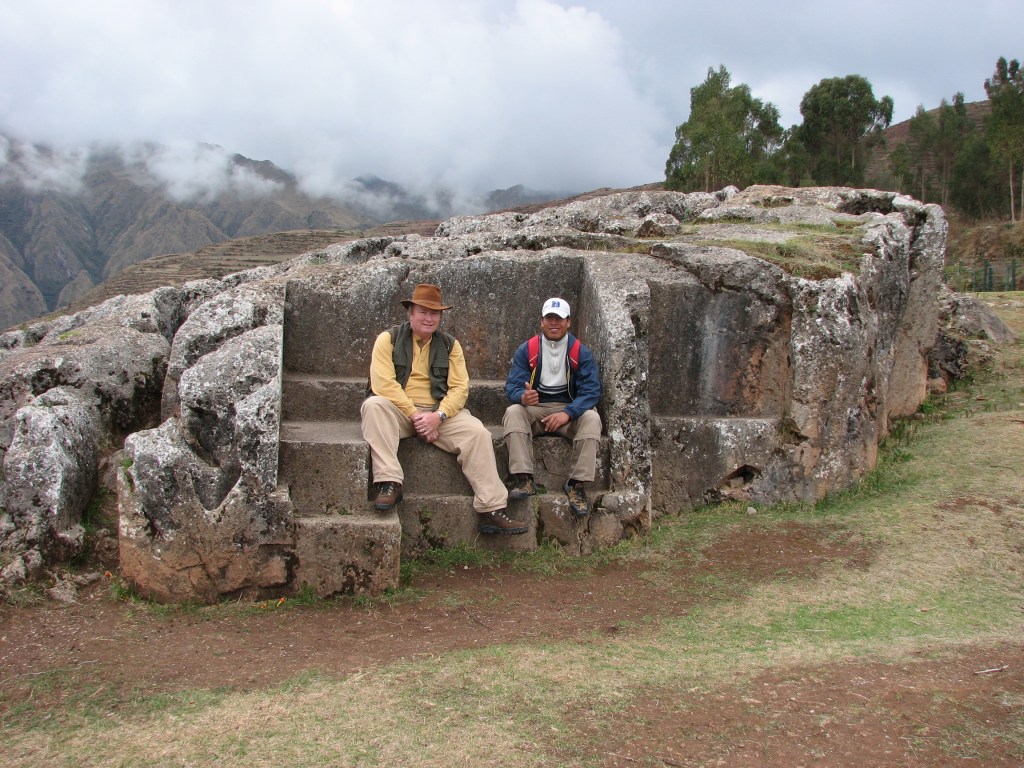 To men sit on carved steps in a huge stone structure at an archaeological site. Clouds hang over mountains in the background.