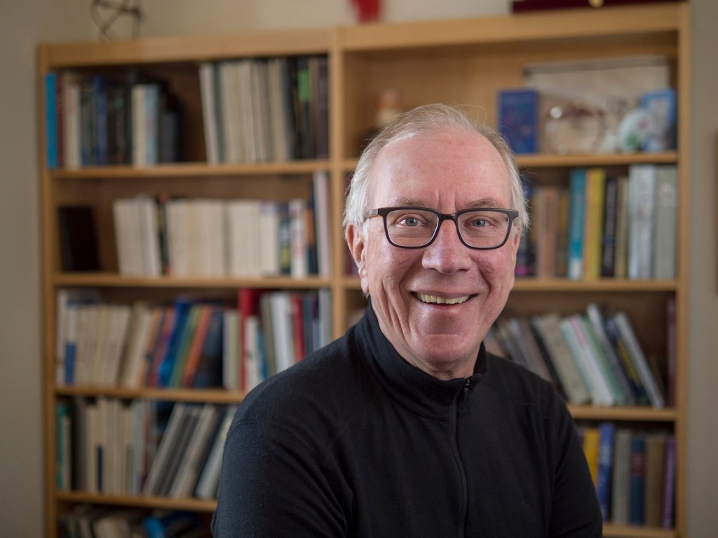 A man wearing glasses smiles at the camera. Behind him is a bookshelf filled with books.