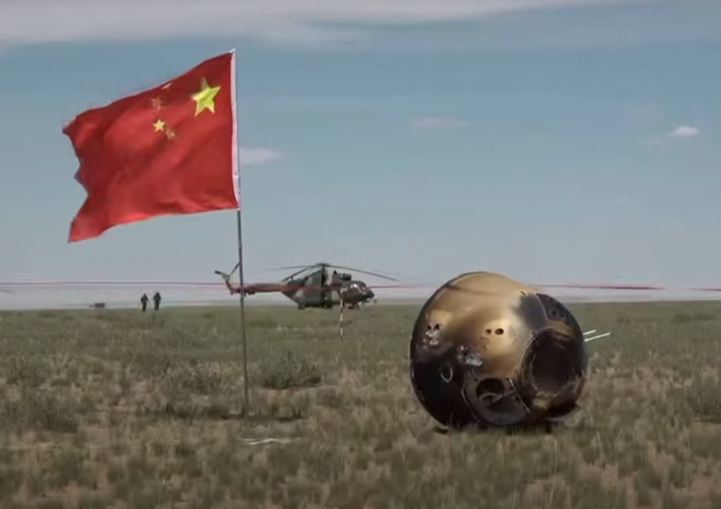 a metal capsule next to a planted Chinese flag on a grassy plain.