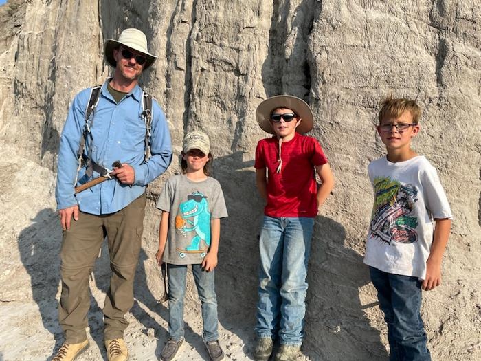 On the far left, an adult man wearing a sun hat. On the right, three young boys. They are all standing in front of a large rock wall outdoors.
