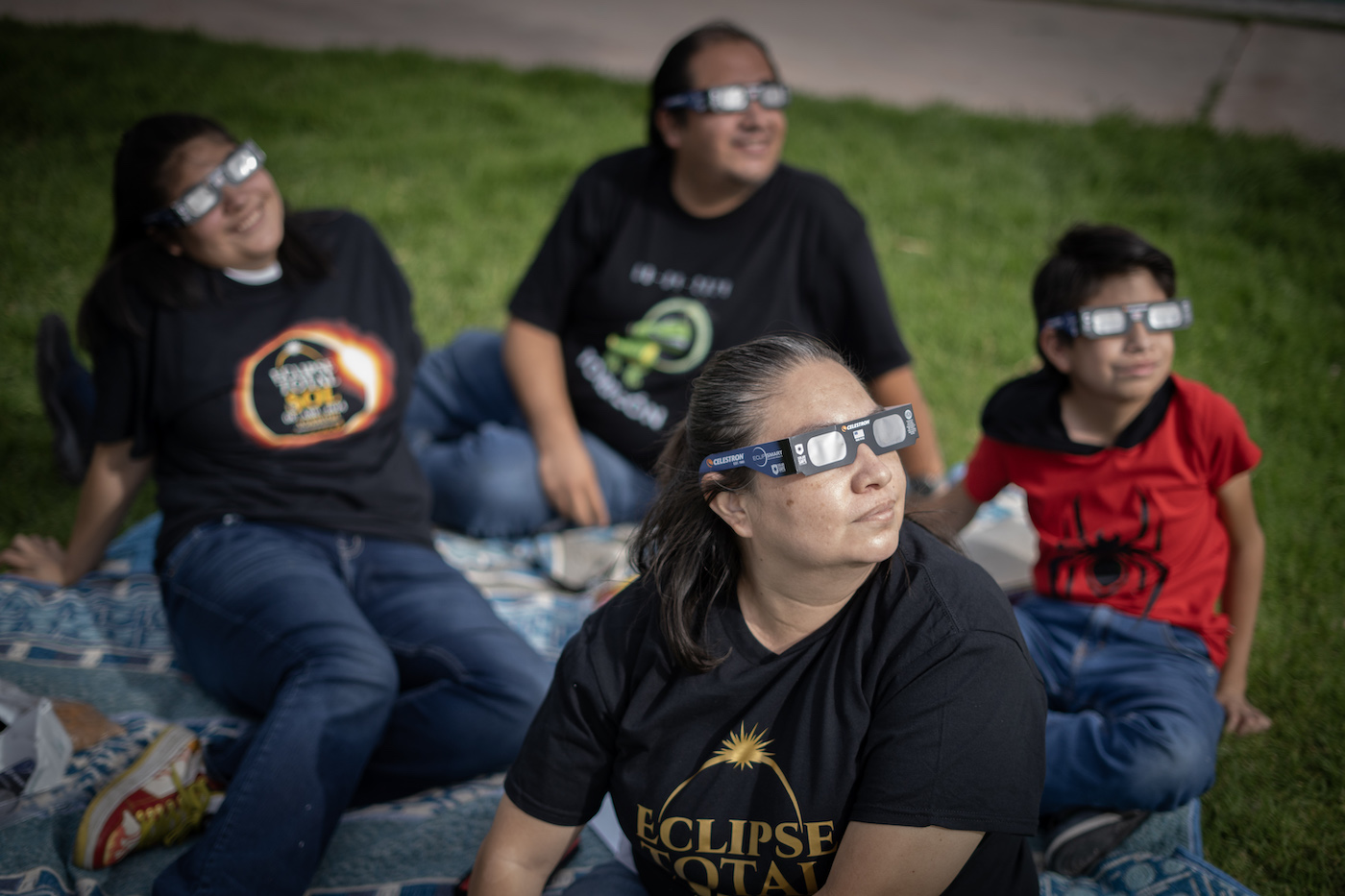 A family of four people wearing eclipse glasses, sitting on a picnic blanket on grass.