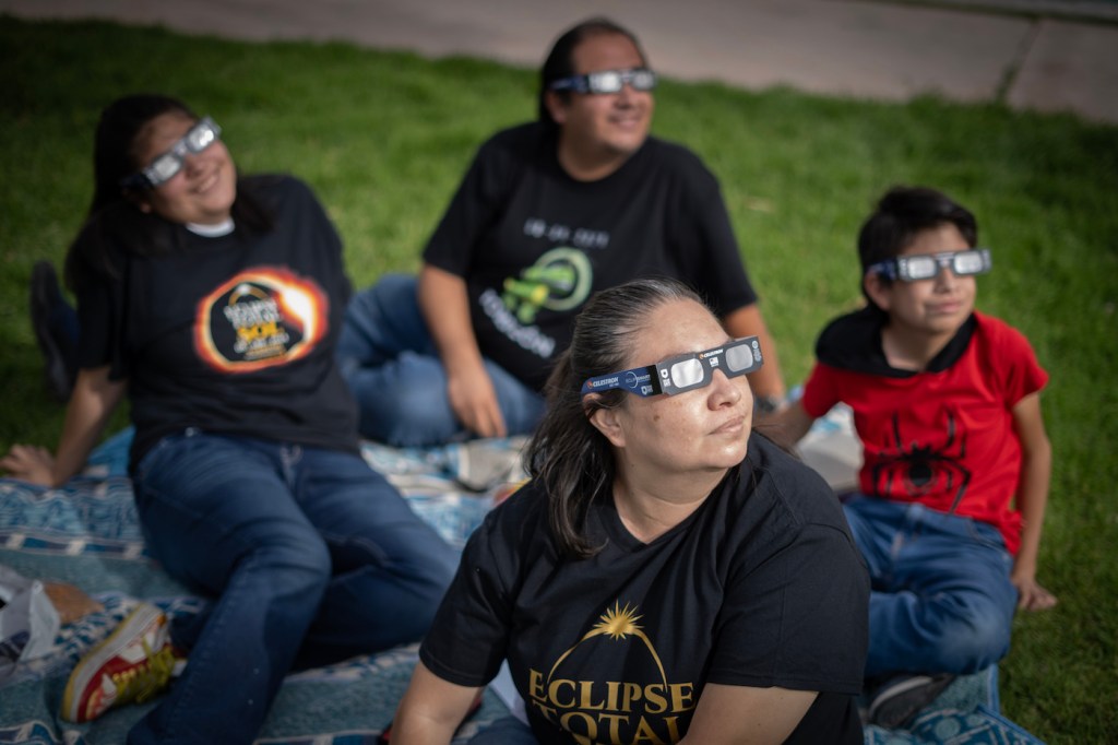A family of four people wearing eclipse glasses, sitting on a picnic blanket on grass.