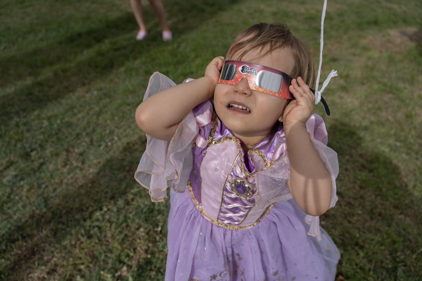 A young girl wearing a princess dress holding eclipse glasses to her eyes, looking at the sky.