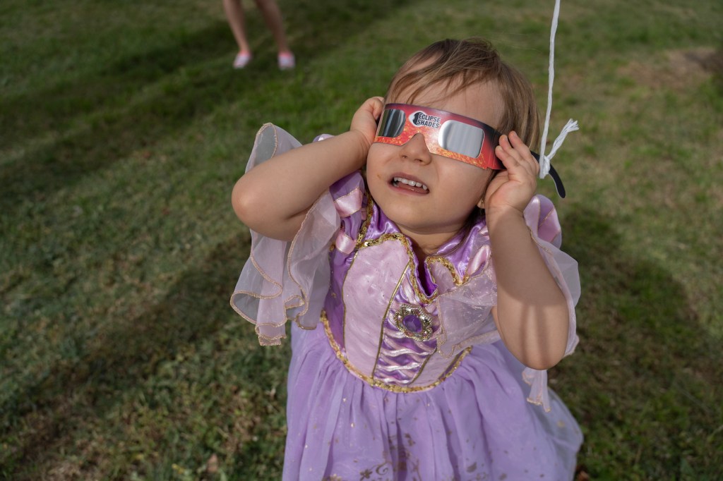 A young girl wearing a princess dress holding eclipse glasses to her eyes, looking at the sky.