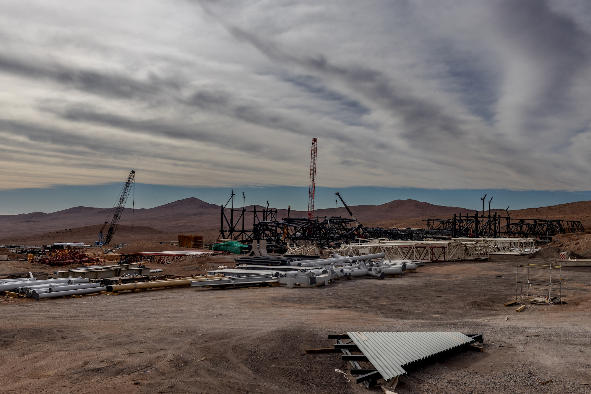 A flat brown landscape with large metal pipes and cranes scattered against the brown desert mountains and cloudy sky.