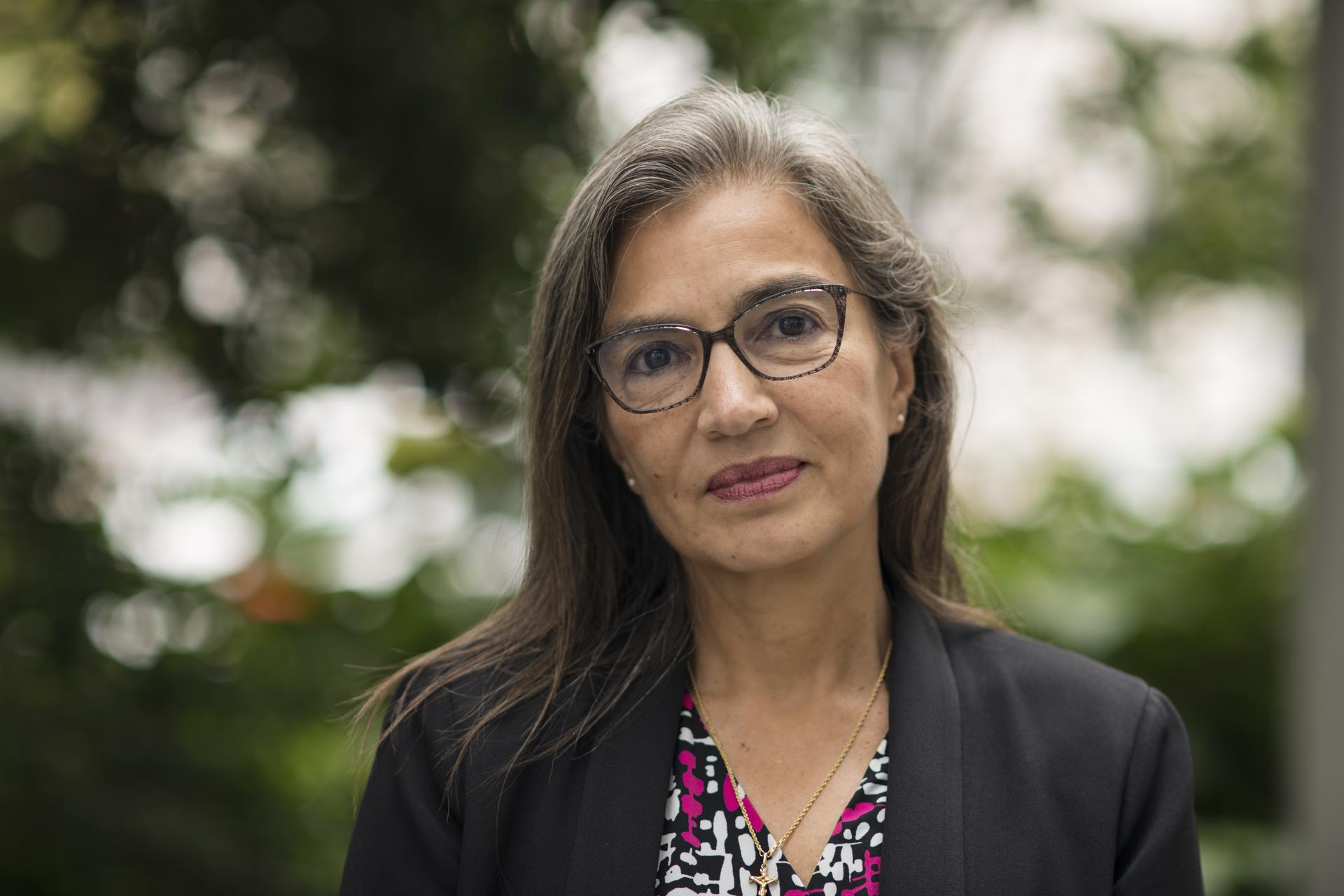 A woman with long hait and glasses looks straight into the camera against a blurry outdoor background