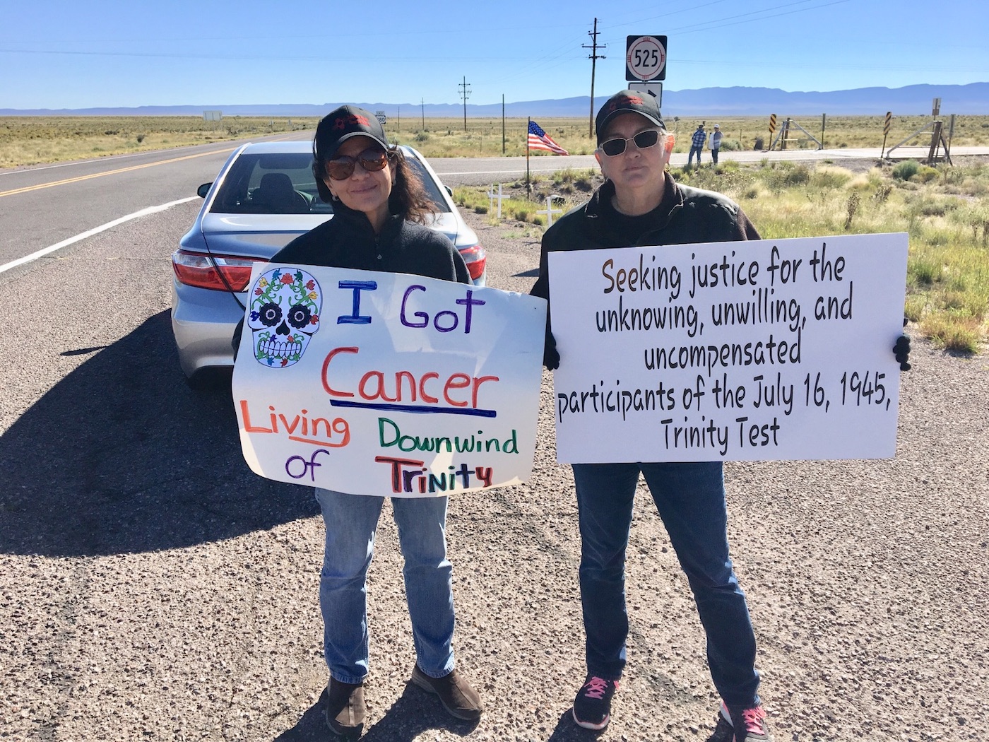 Two people holding signs. One says: "I got cancer living downwind of Trinity" and the other says: "Seeking justice for the unkniwing, unwilling, and uncompensated participants of the July 16, 1945 Trinity Test"
