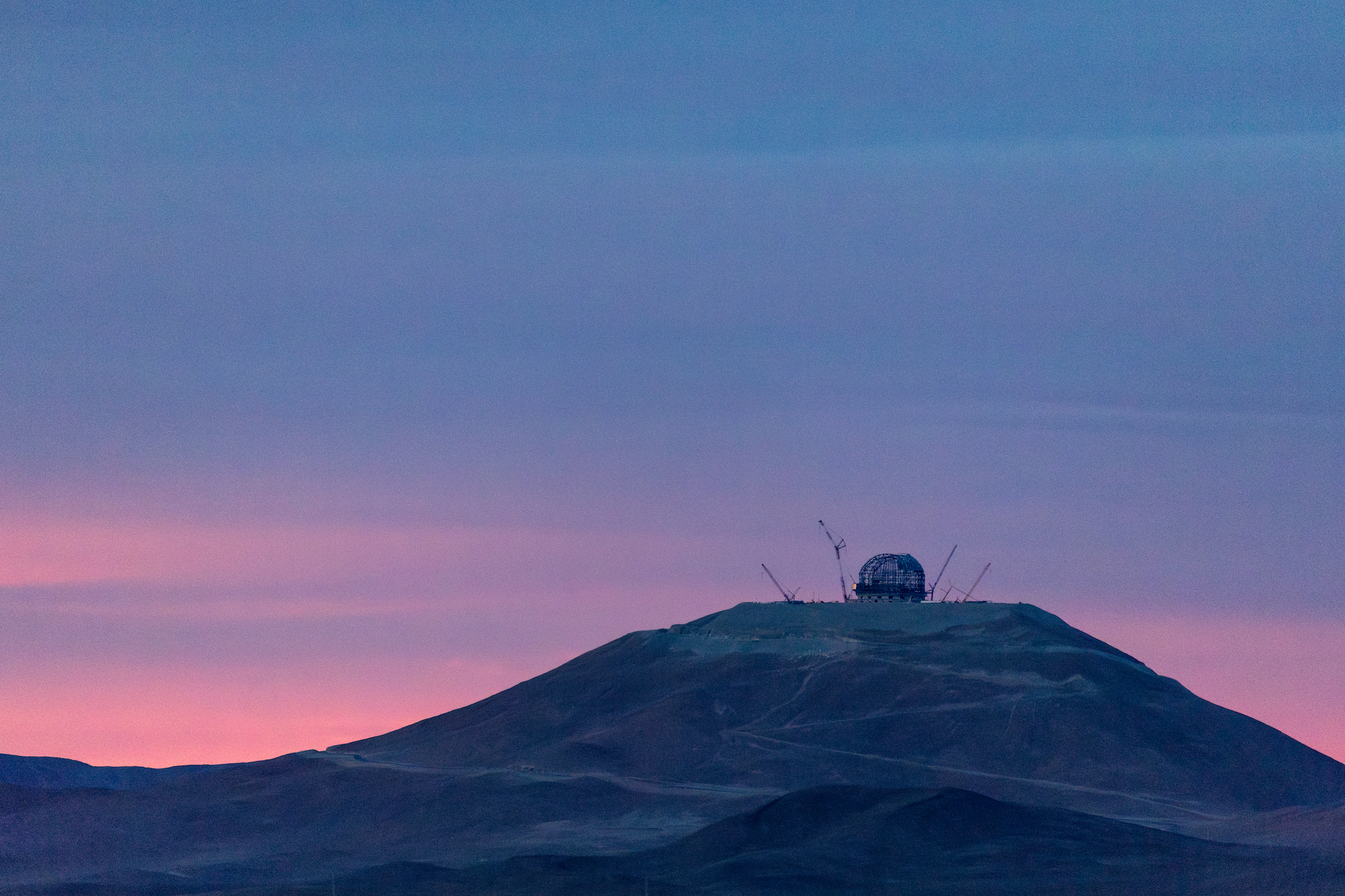 A barren distant mountain peak with a huge dome and many cranes around it was silhouetted against the soft blue and pink sky.