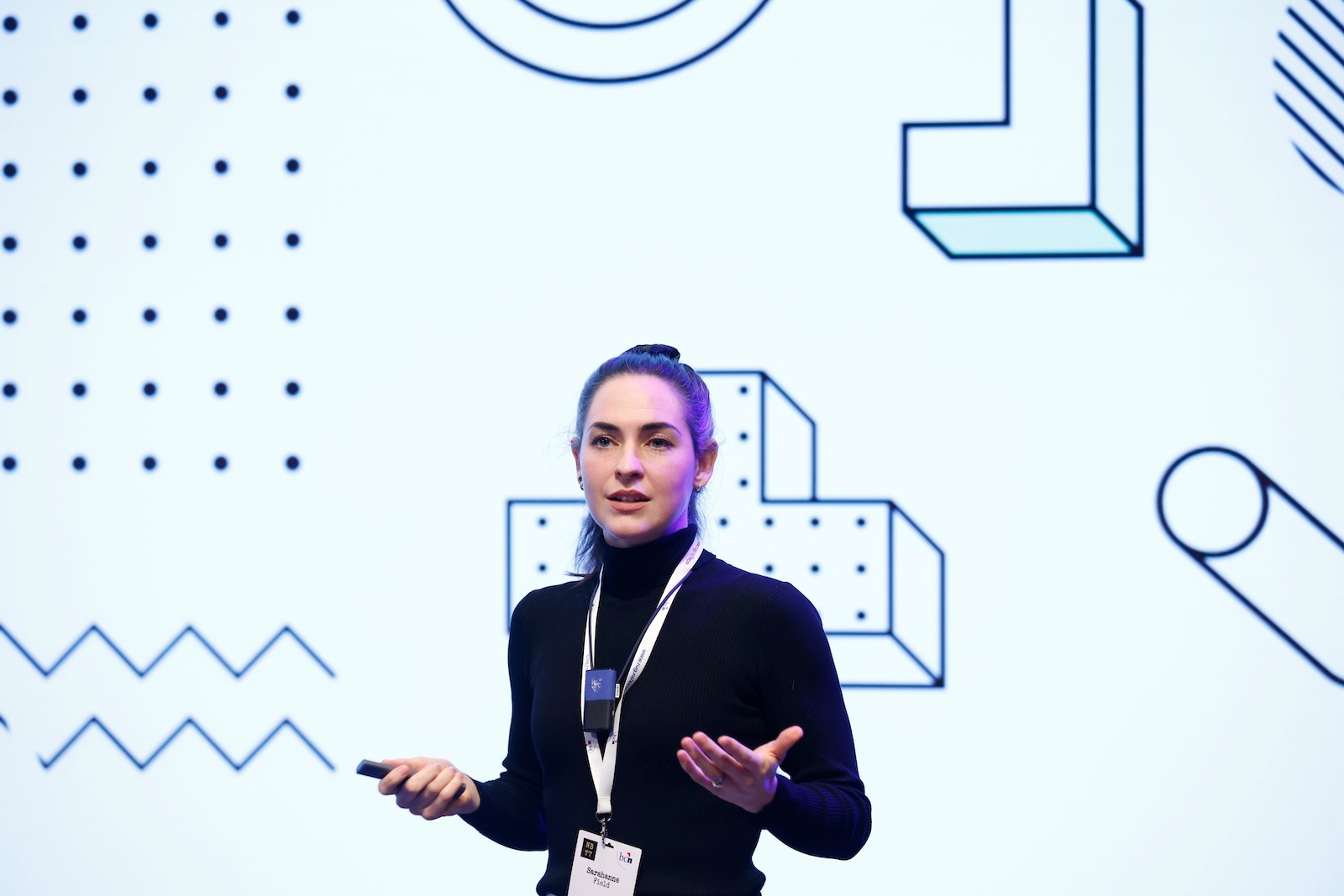 a white woman wearing a black turtleneck wearing a lanyard and a necklace mic gesturing with her hands speaking on a stage with abstract graphics behind her