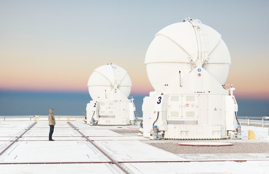 A woman in a coat stands looking up at two gigantic white spherical telescopes on a white platform against a pale blue twilight.