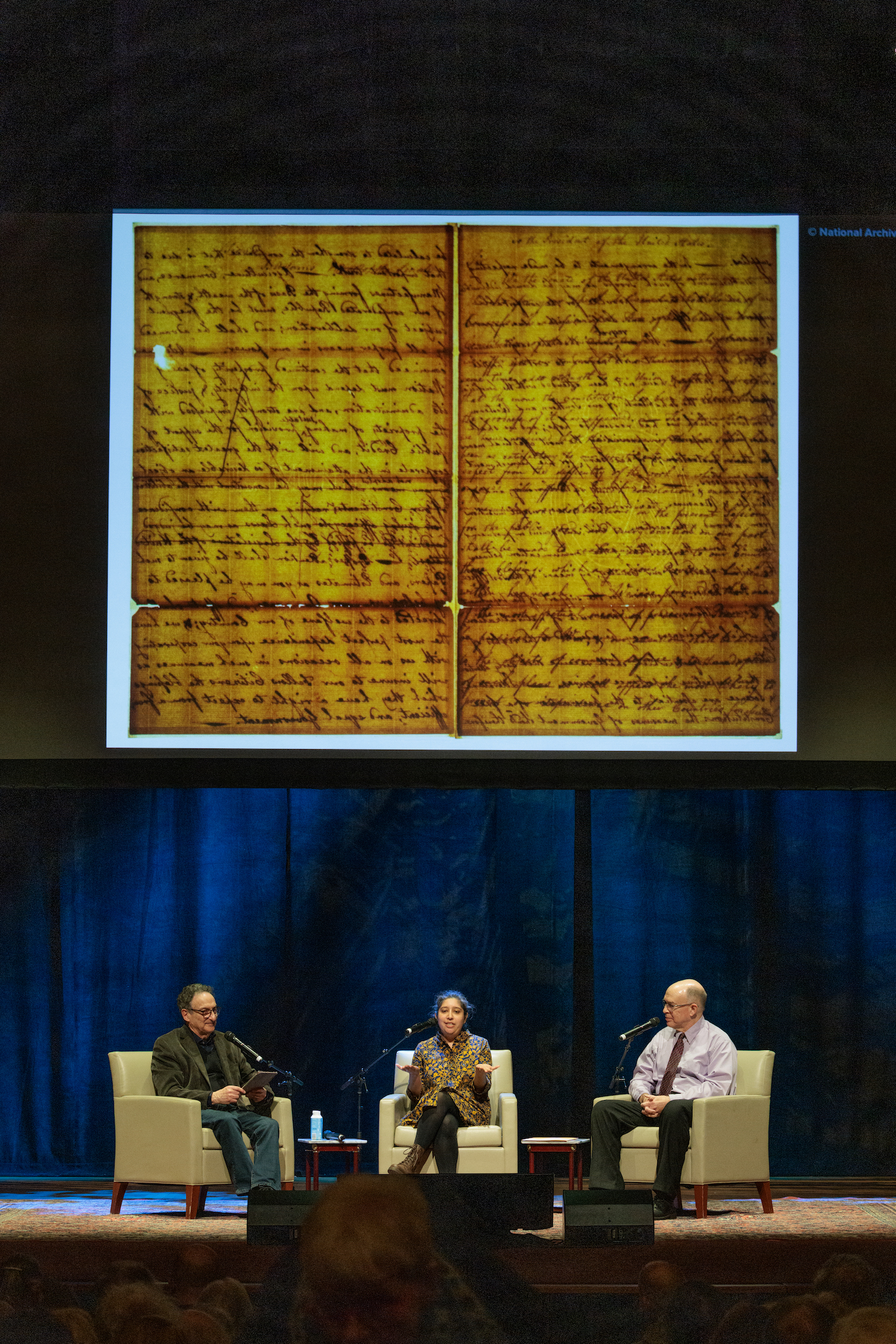 A yellowed document projected above a group of three people on a stage