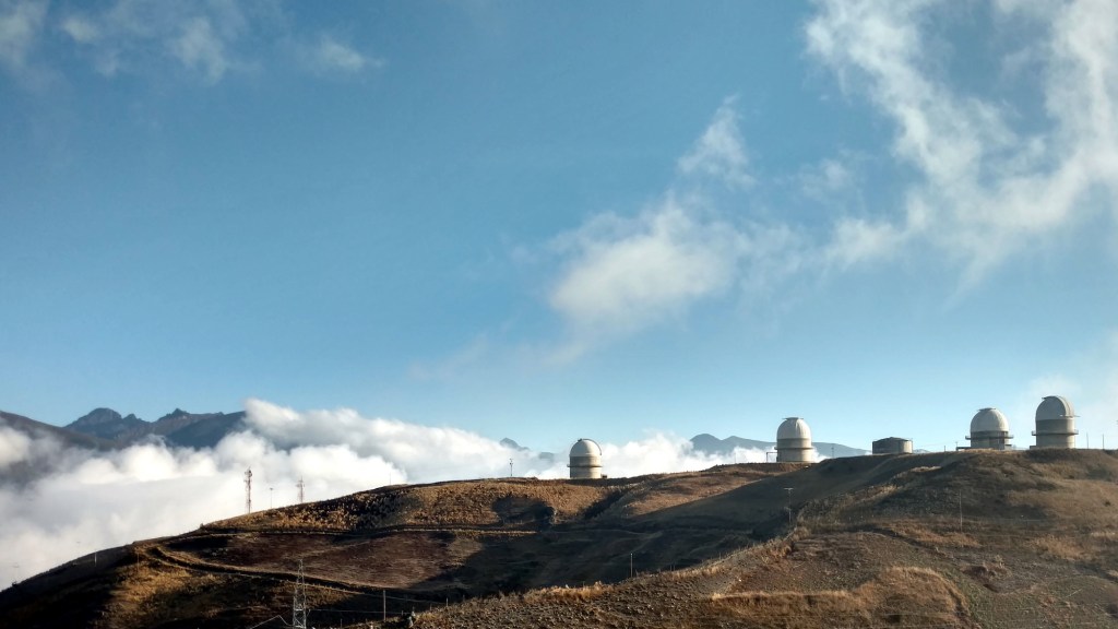 View of hills with four large telescope domes on a ridge against a blue sky with clouds.