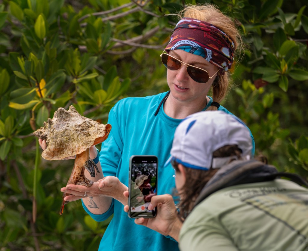 A woman holds a conch shell with the animal drooping out of it, as another person takes an iPhone photo of the conch.