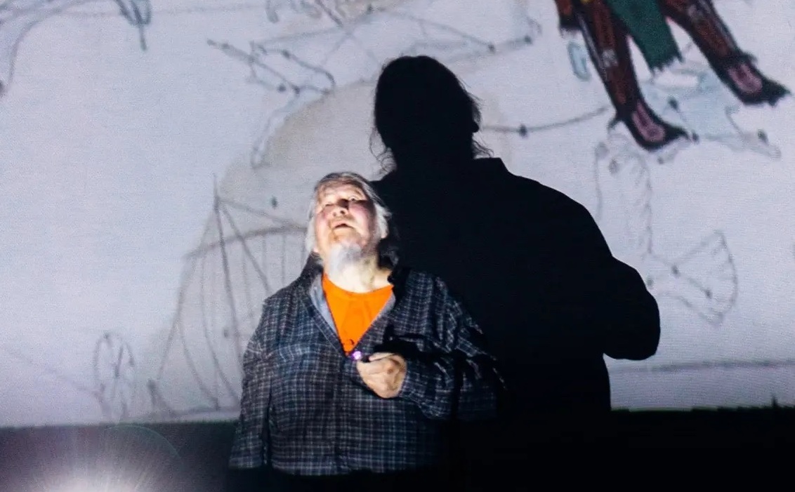 a man in a domed room looking up at a projected star map of indigenous constellations