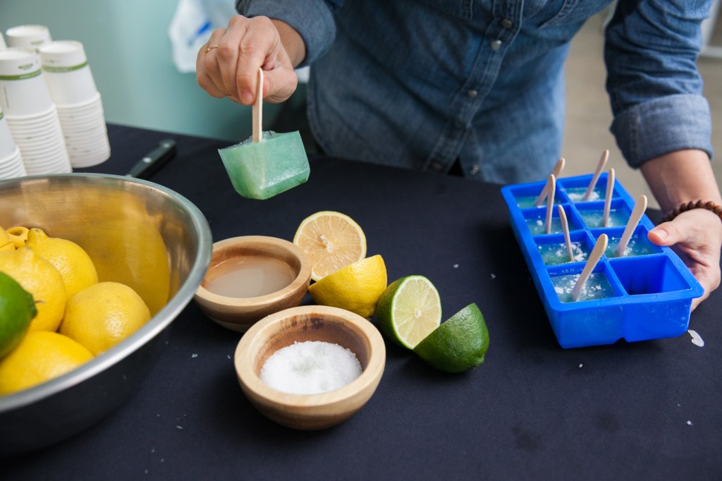 Close -up of a table where a woman holds a greenish cubic popsicle in one hand and a tray of more of them in another, amid bowls of lemons, limes, and salt.