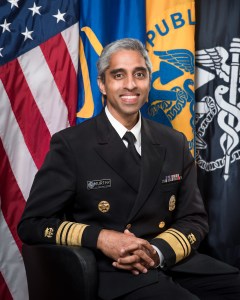 A man wearing an official US uniform in front of 4 flags, including the American flag.