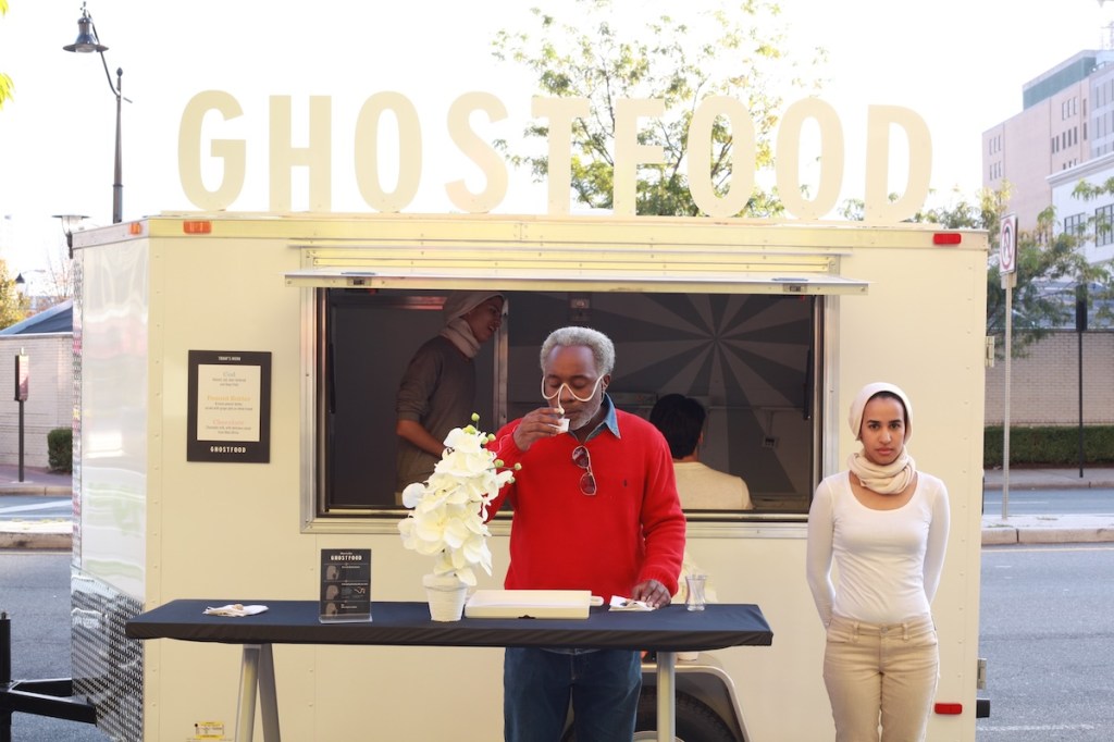 A black man in a red sweater sips from a cup while inhaling from a wired wearable facial device, while standing in front of a white food truck.