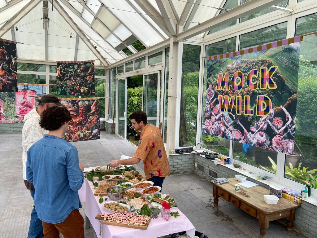 In a greenhouse-like building with glass walls, a man serves two people food from a table with colorful trays of food.