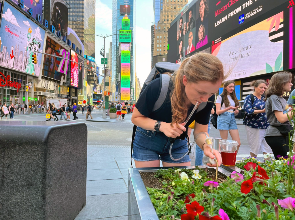 A white woman bends down over a flower planter with one end of a tube in her mouth and the other end in the dirt. Times Square buildings and ads tower behind her.