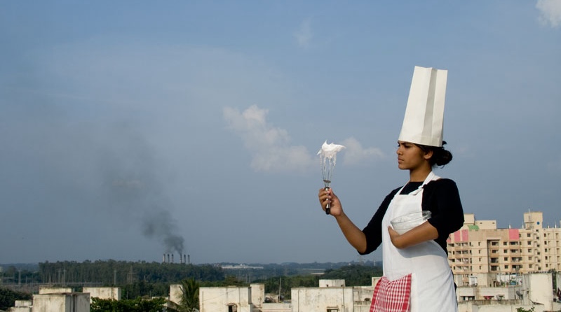 A woman wearing a chef's hat hols a whisk with white foam on it in front of her. In the background, smokestacks belch smog.