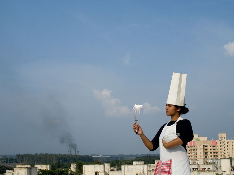 A woman wearing a chef's hat hols a whisk with white foam on it in front of her. In the background, smokestacks belch smog.