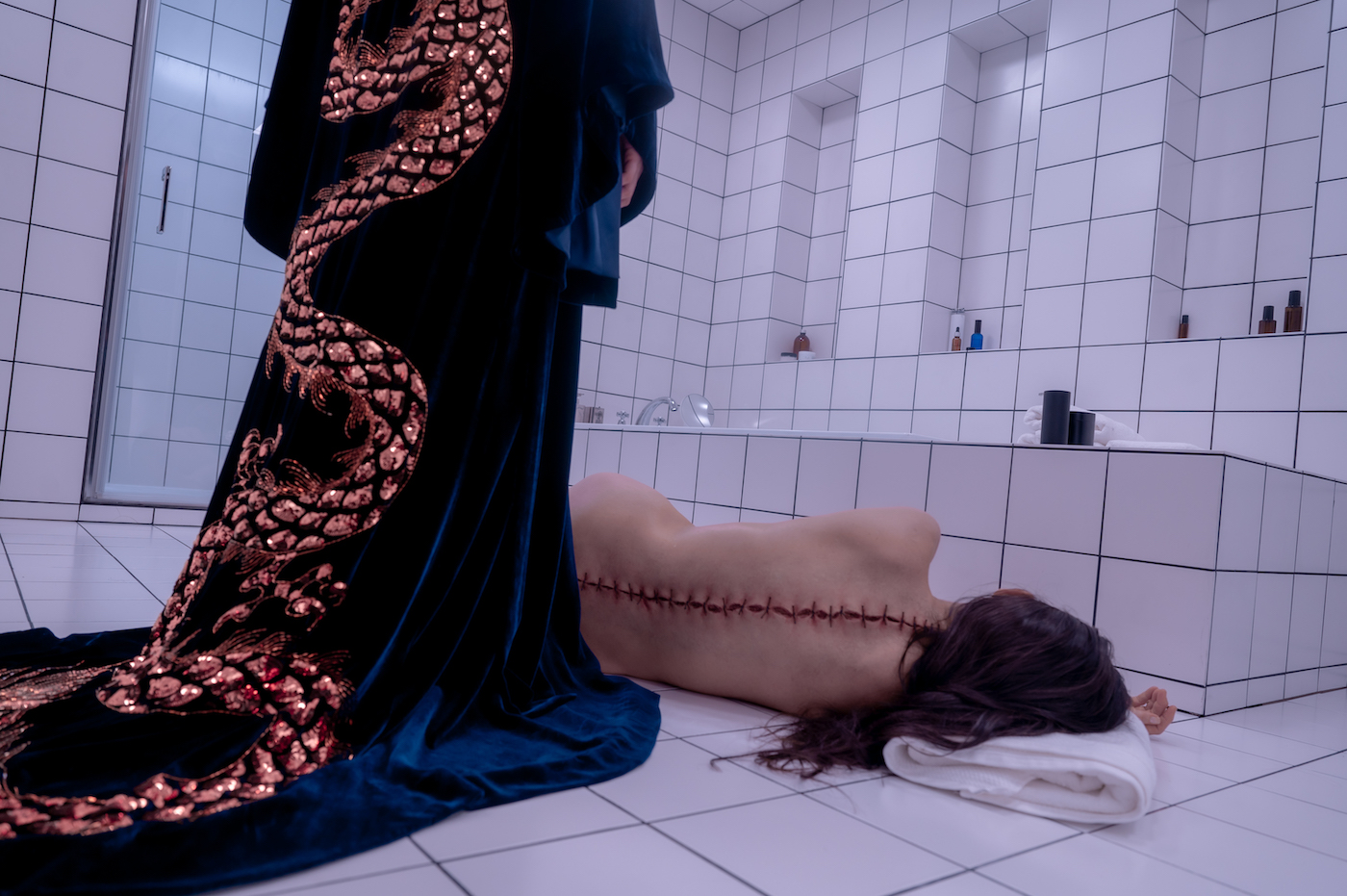 A woman with a wound all down her spine lying naked on a bathroom floor. Someone dressed in a robe stands over her.