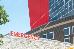 The illuminated red letters spell emergency and is affixed to white metal beams. There are windows in the background of a large hospital building.