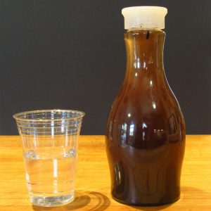 A bottle filled with dark-colored liquid stands on a wooden surface against a dark background next to a clear plastic cup containing clean water.