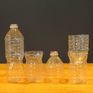 Several disposable plastic water bottles or pieces of plastic bottles are arranged on a wooden surface against a dark background. The first bottle on the left is whole. In the center, a bottle is cut in half. On the right, the top of the cut bottle has been inserted into the bottom of the cut bottle to create a filter.