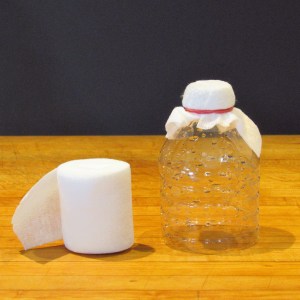 The top of a disposable plastic water bottle sits on a wooden surface against a dark background. Its top, or spout, is covered with a piece of white gauze secured by a rubber band. Next to the bottle is a roll of gauze.