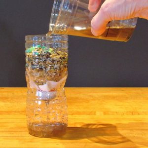 A hand pours a brownish liquid from a clear plastic cup into a plastic bottle filled with layers of various materials, including colorful pebbles and grains. The bottle sits on a wooden surface against a dark background.