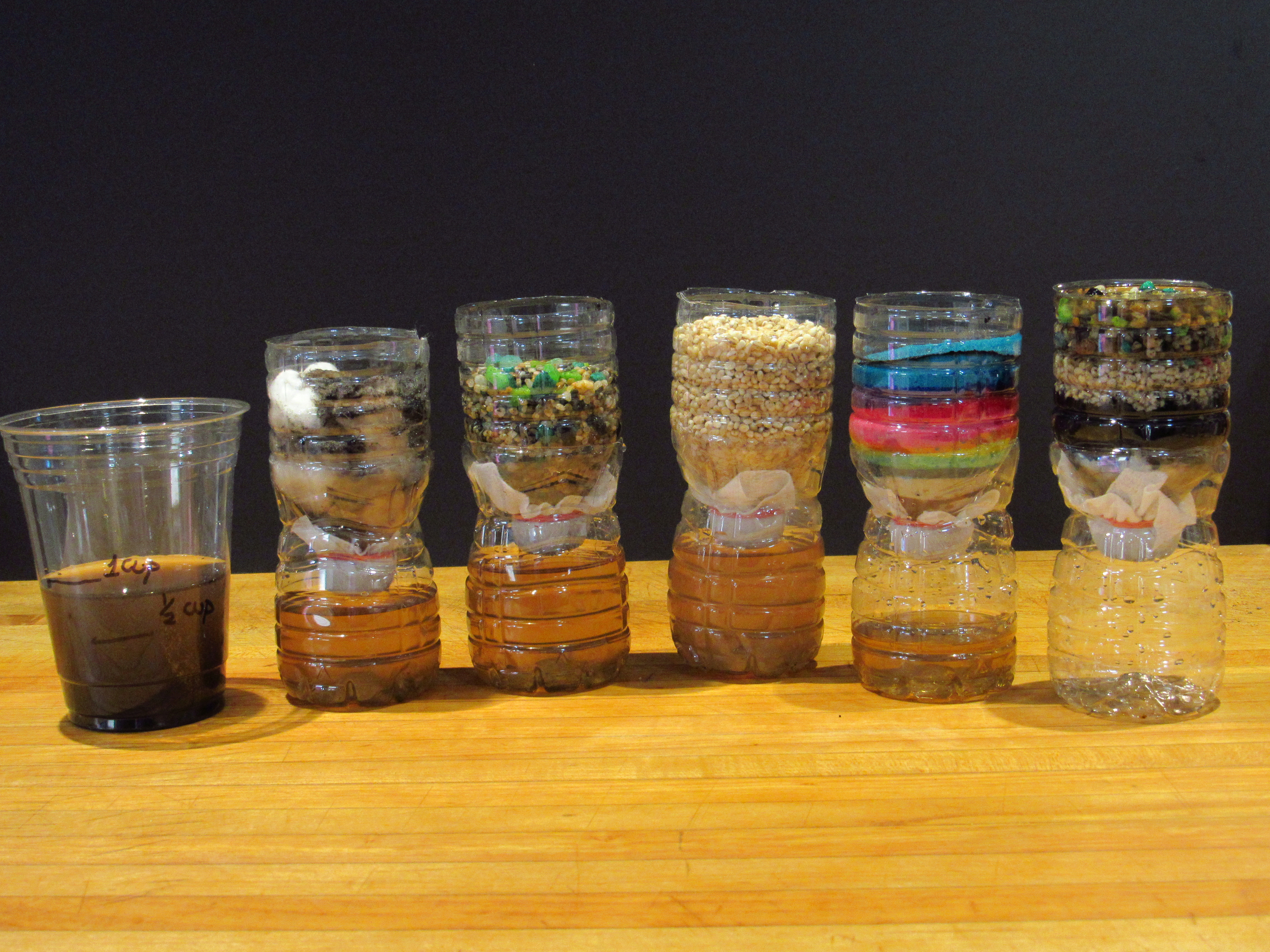 Five clear plastic bottles are arranged in a row on a wooden surface against a dark background. Each bottle contains different materials such as colored sands, grains, and pebbles. A clear plastic cup with dark liquid is placed at the far left.