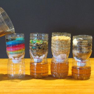 Four clear plastic bottles sit on a wooden surface against a dark background. Each bottle contains different types of filtering media, including colored sand, grains, and pebbles. A brownish liquid is being poured into the leftmost bottle, which has rainbow-colored layers.