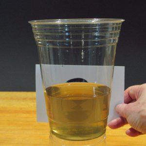 A clear plastic cup filled with brownish-yellow water sits on a wooden surface with a dark background. A hand holds a white card on which a black and white circular object is drawn behind the cup. The object cannot be seen clearly through the liquid.