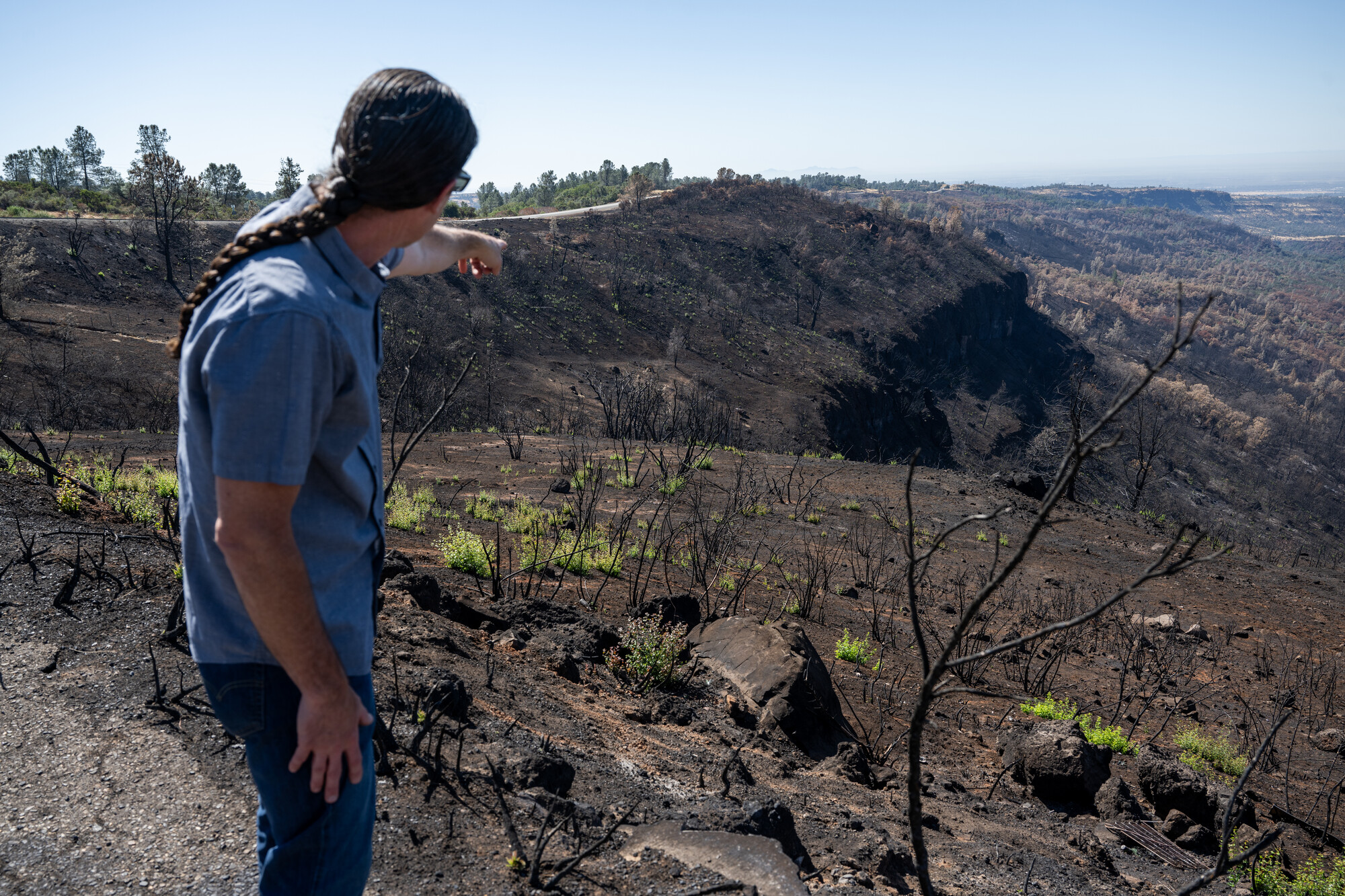 A man standing on a low mountain, pointing at the other side of it. The landscape is burnt and ashy.