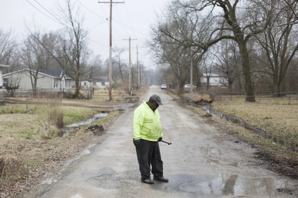 A Black man stands on an empty dirt road looking down at water bubbling from a drain, on an overcast winter day