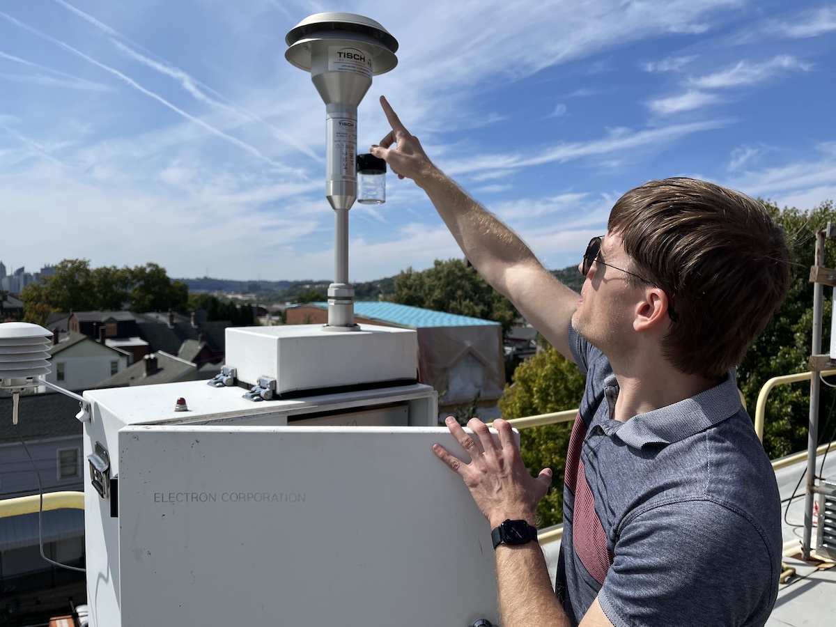 A man on a rooftop points to the round top of a boxy device, against a blue sky in bright sun