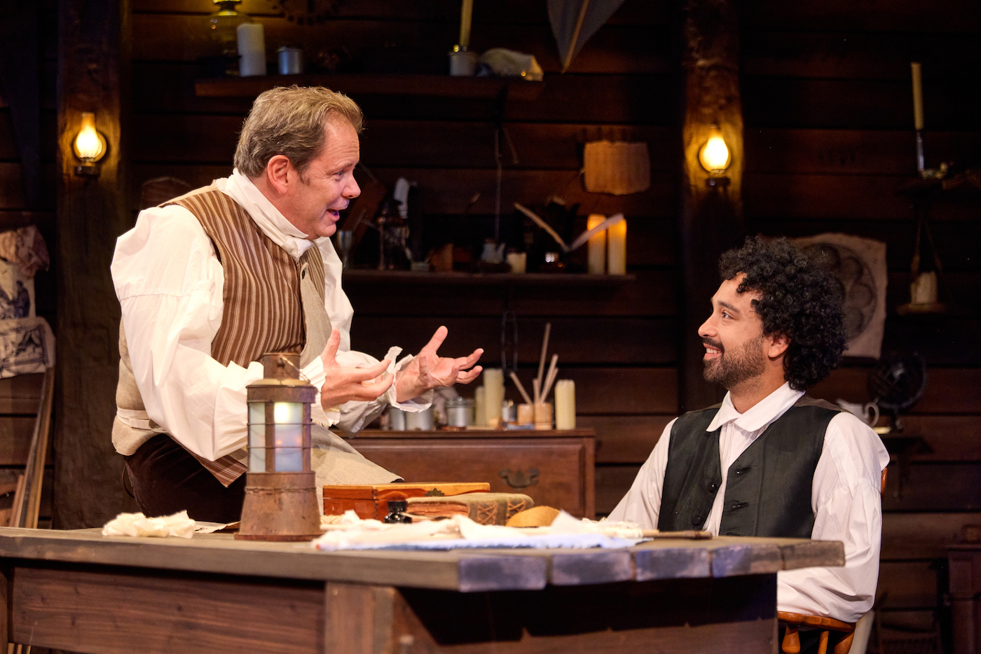 Two actors in costume sitting at a table on stage.