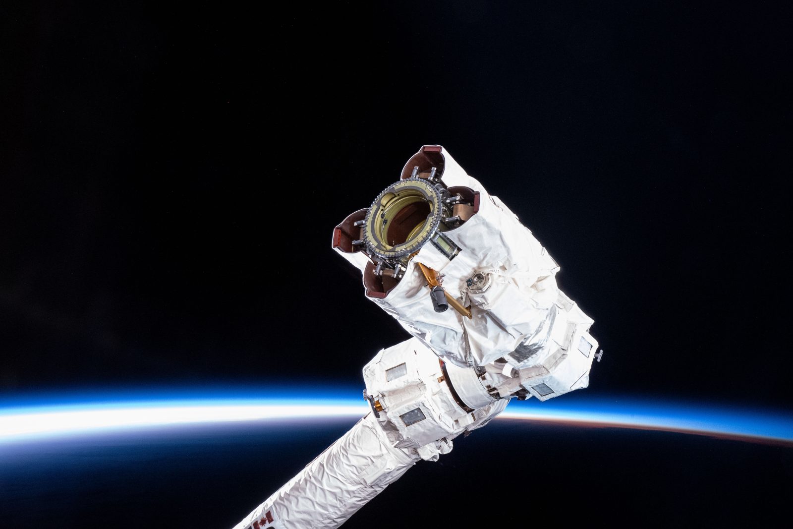 A robotic arm extends from the ISS with Earth’s horizon glowing faintly against the darkness of space. The arm’s end, with a central ring of latches to form a specialized grappling mechanism, is shown in close-up.