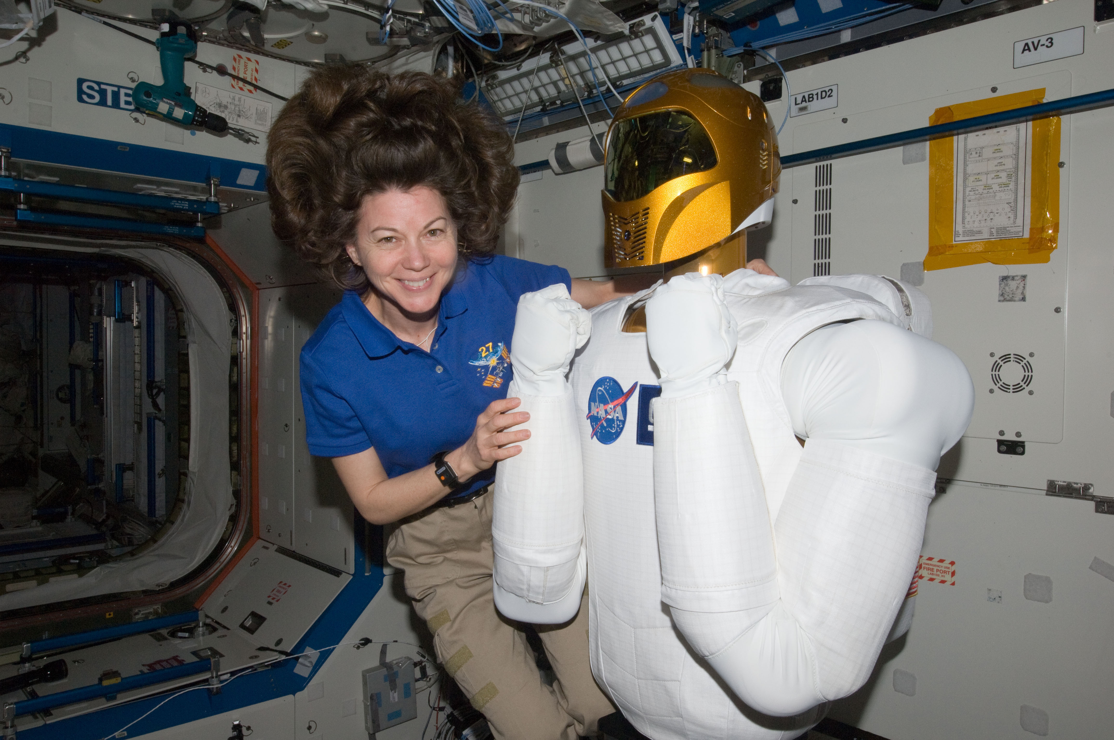 An astronaut aboard the International Space Station poses with Robonaut 2, a humanoid robot, while her hair floats in zero gravity. The background shows the space station’s interior with equipment and a NASA logo visible.