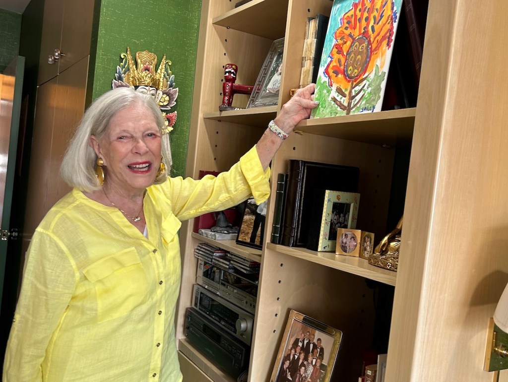 An older woman looking happy and healthy, reaching up at a painting she made on a shelf.