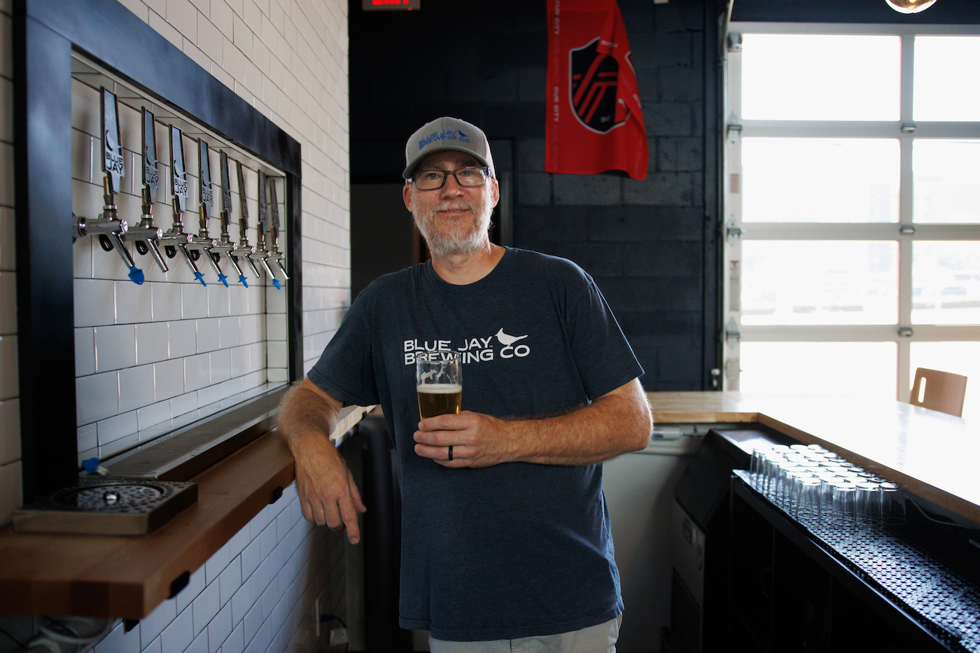 A man wearing glasses and a baseball cap holds a beer.