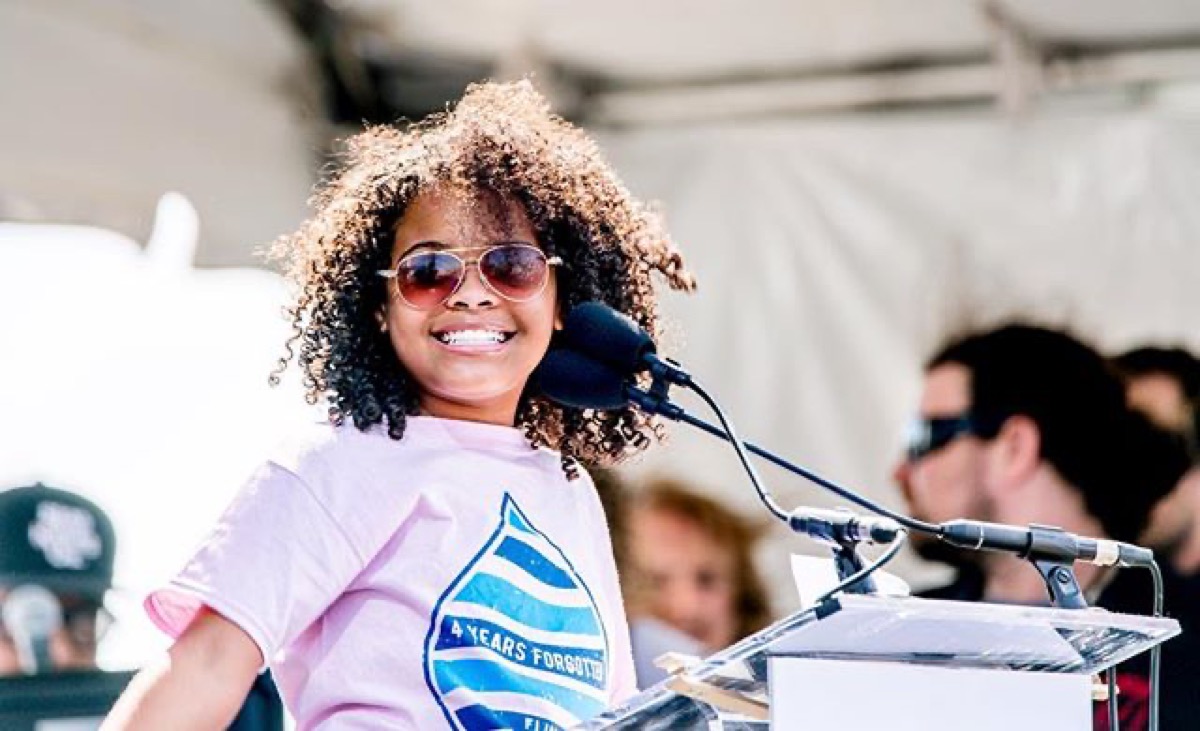 A young girl with curly hair smiles brightly while standing at a podium, speaking into a microphone. She wears sunglasses and a light pink T-shirt with a blue water drop design on the front that reads 4 years forgotten.