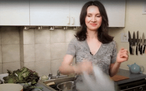 A woman in a kitchen spins a bundle of lettuce inside a white plastic bag. She smiles as she swings the bag in a circular motion, using the motion to dry the leaves. The kitchen background shows a kettle, a row of knives, a kitchen sink, and cabinets.